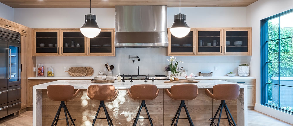 A modern kitchen featuring wooden cabinets and ceiling, a white countertop with three brown leather stools, stainless steel appliances, and pendant lighting. The space is illuminated by natural light from a large window with a view of greenery outside. Decorative items, including a vase with flowers and some fruits, are placed on the countertop.