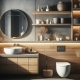 a rustic-modern bathroom interior design with a sleek wooden aesthetic. The color palette consists of grays and wood, with contrasting textures like matte walls and glossy cabinets. Accent lighting under the mirror, shelves, and cabinet edges provides a warm glow. The bathroom features a round countertop basin, a glass-enclosed shower area, a wall-mounted toilet, and built-in shelves stocked with towels and toiletries. Various decorative elements such as dried plants in vases, small potted plants, and woven baskets add a natural touch to the space. The light from a frosted glass window creates a bright spot.