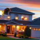 A well-lit suburban home at dusk showcasing various home security features AT CLOSE-UP. Visible security cameras are mounted near the front door and along the roofline. Motion-sensor lights illuminate the front yard and driveway.
