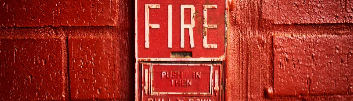 A close-up of a vintage red fire alarm mounted on a textured brick wall. The alarm features the word 'FIRE' in large, bold white letters, with instructions below that read 'PUSH IN THEN PULL DOWN.' The surrounding brick wall is painted in a matching red shade, giving the image a monochromatic and intense appearance.