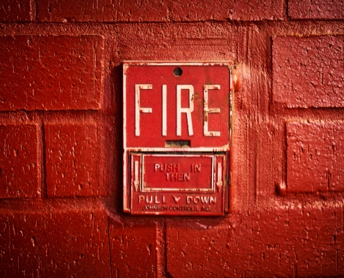 A close-up of a vintage red fire alarm mounted on a textured brick wall. The alarm features the word 'FIRE' in large, bold white letters, with instructions below that read 'PUSH IN THEN PULL DOWN.' The surrounding brick wall is painted in a matching red shade, giving the image a monochromatic and intense appearance.