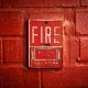 A close-up of a vintage red fire alarm mounted on a textured brick wall. The alarm features the word 'FIRE' in large, bold white letters, with instructions below that read 'PUSH IN THEN PULL DOWN.' The surrounding brick wall is painted in a matching red shade, giving the image a monochromatic and intense appearance.