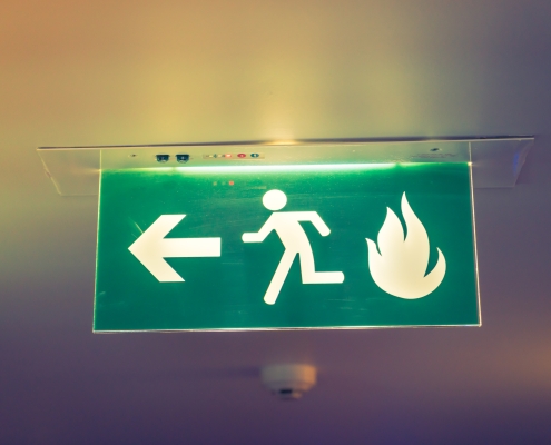 A green emergency exit sign mounted on the ceiling, featuring a white icon of a running person and an arrow pointing left towards the exit. A flame symbol is also present, indicating the direction for safe evacuation in case of a fire. The sign is illuminated, enhancing its visibility in low-light conditions.