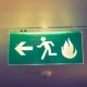 A green emergency exit sign mounted on the ceiling, featuring a white icon of a running person and an arrow pointing left towards the exit. A flame symbol is also present, indicating the direction for safe evacuation in case of a fire. The sign is illuminated, enhancing its visibility in low-light conditions.