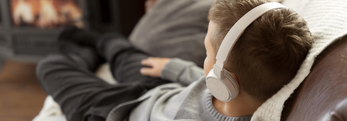 A young child wearing white headphones, reclining comfortably on a sofa with a cozy blanket. The child is facing away from the camera, looking towards a fireplace in the background. The warm, softly lit scene suggests relaxation and comfort in a home environment, with the glow of the fire adding to the cozy atmosphere.